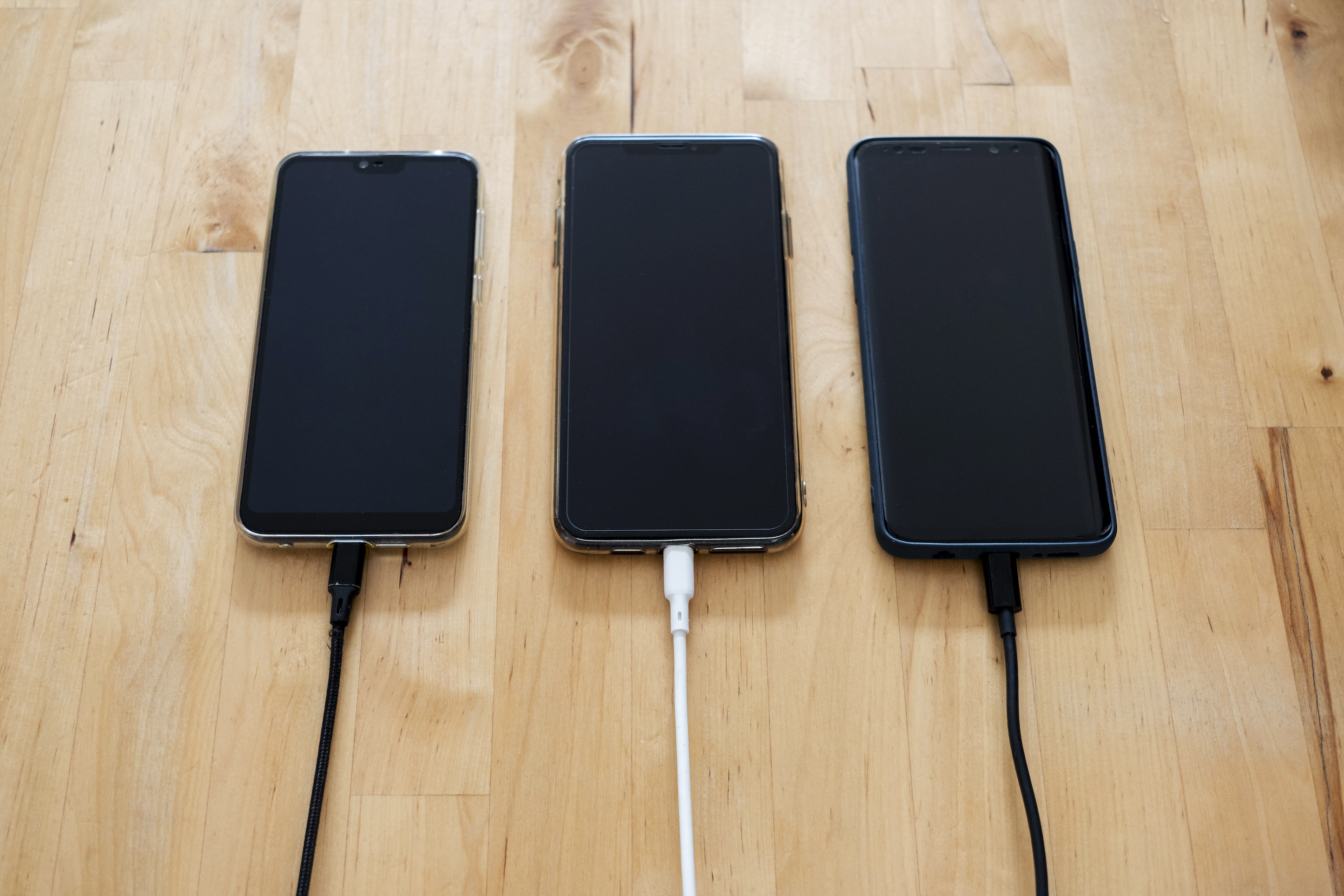 Trois téléphones qui sont alignés en train de chargeur avec 2 câbles de rechargement noir et 1 blanc sur une table en bois clair