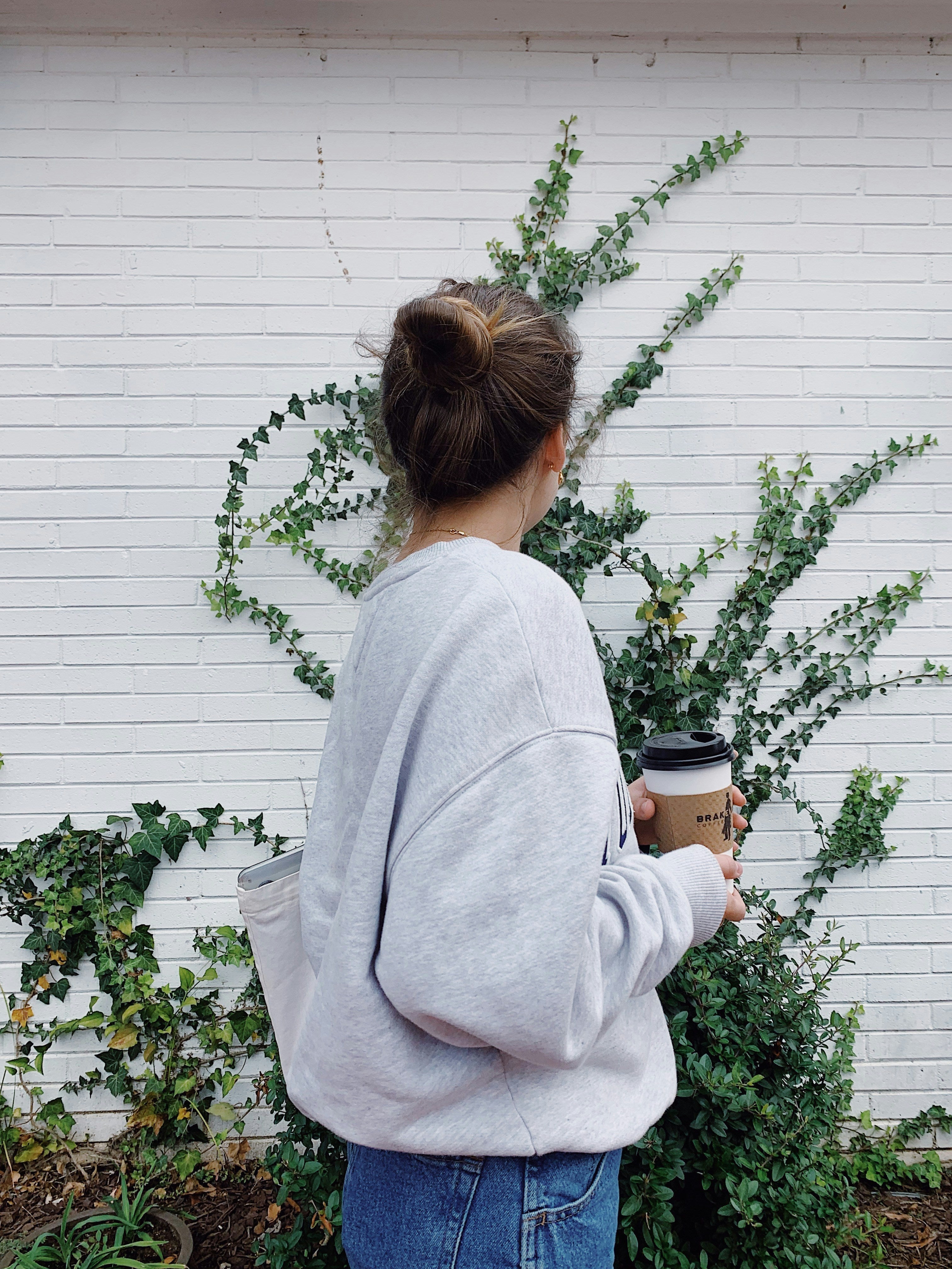 sweat gris sans capuche - femme - elle porte avec un jean - la photo à été prise devant une maison avec un arbuste avec un café à la main