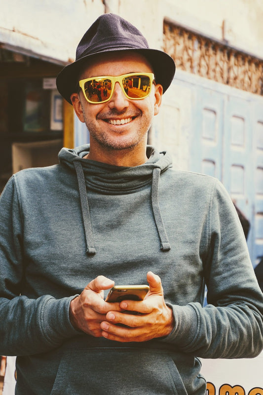 Homme avec sourire qui a un téléphone, sweat gris, lunettes de soleil et chapeau violet