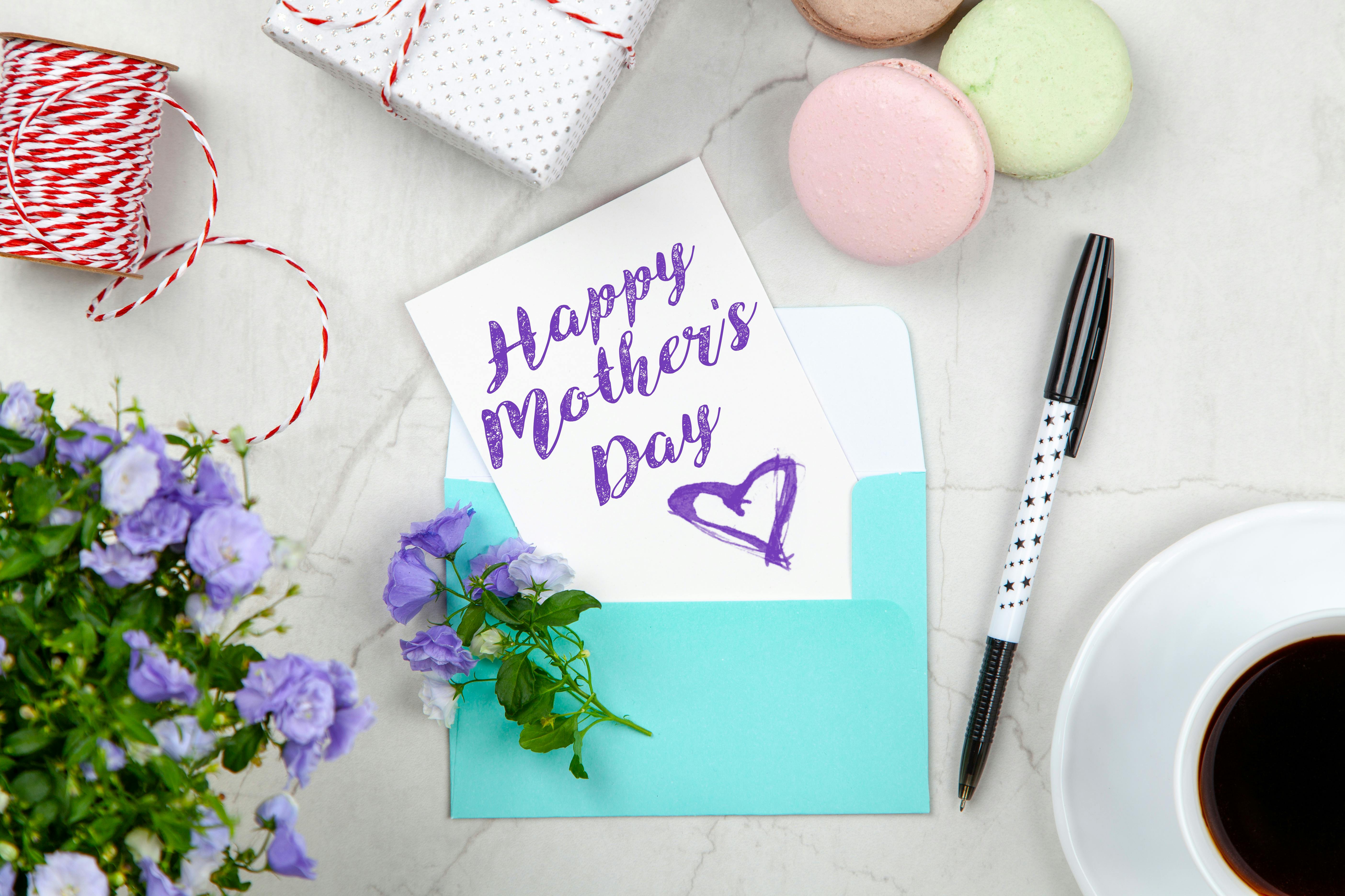 feuille avec écrit "happy mothers day" et un coeur en violet avec une enveloppe bleu , des macarons rose vert et marron pale, un  cadeau et du fil rouge et blanc. Il y a un café et des fleurs violettes sur le côté
