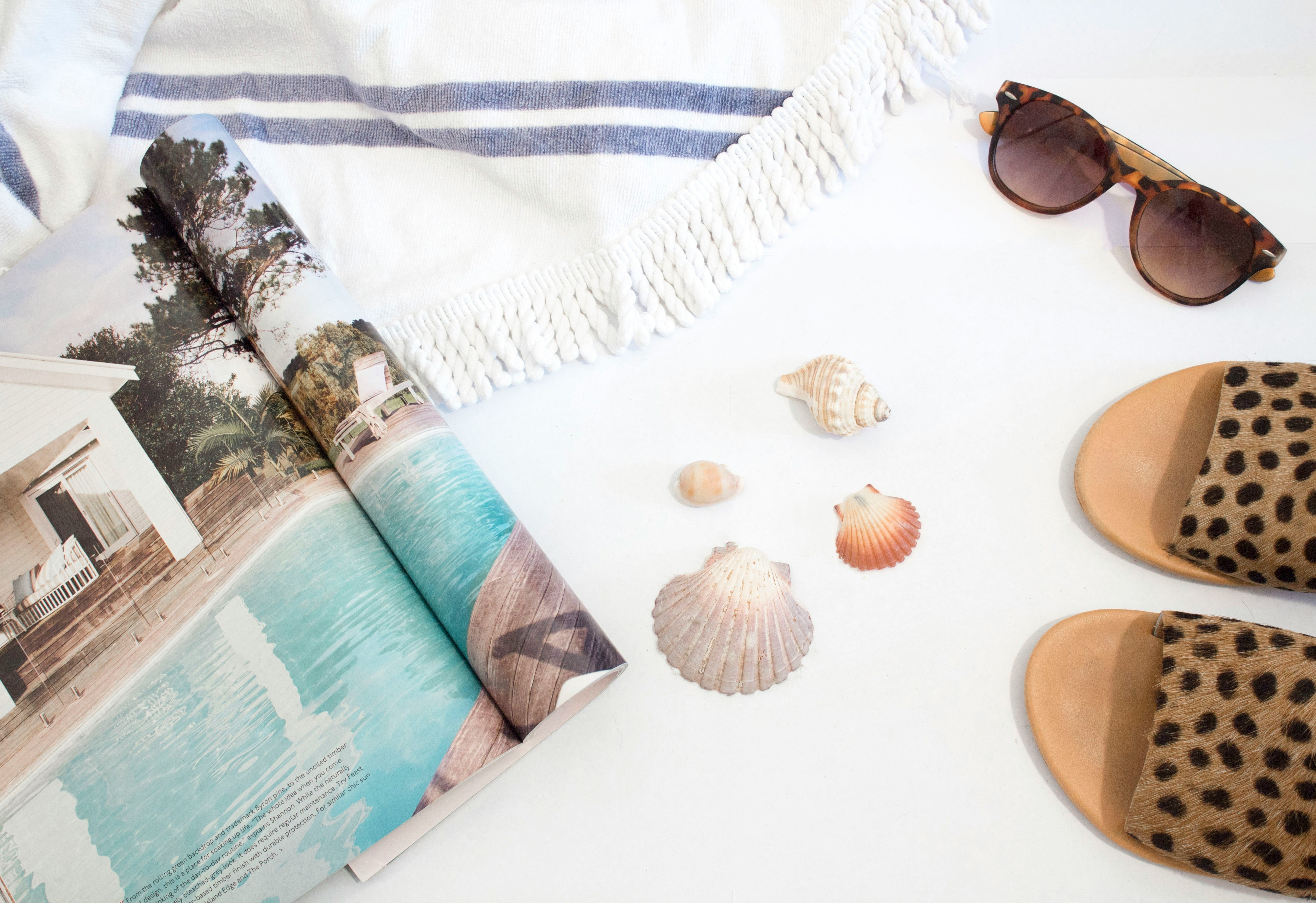 lunettes de soleil, coquillages, claquettes à motifs léopard, un magasine et une serviette de plage beige sur un fond blanc
