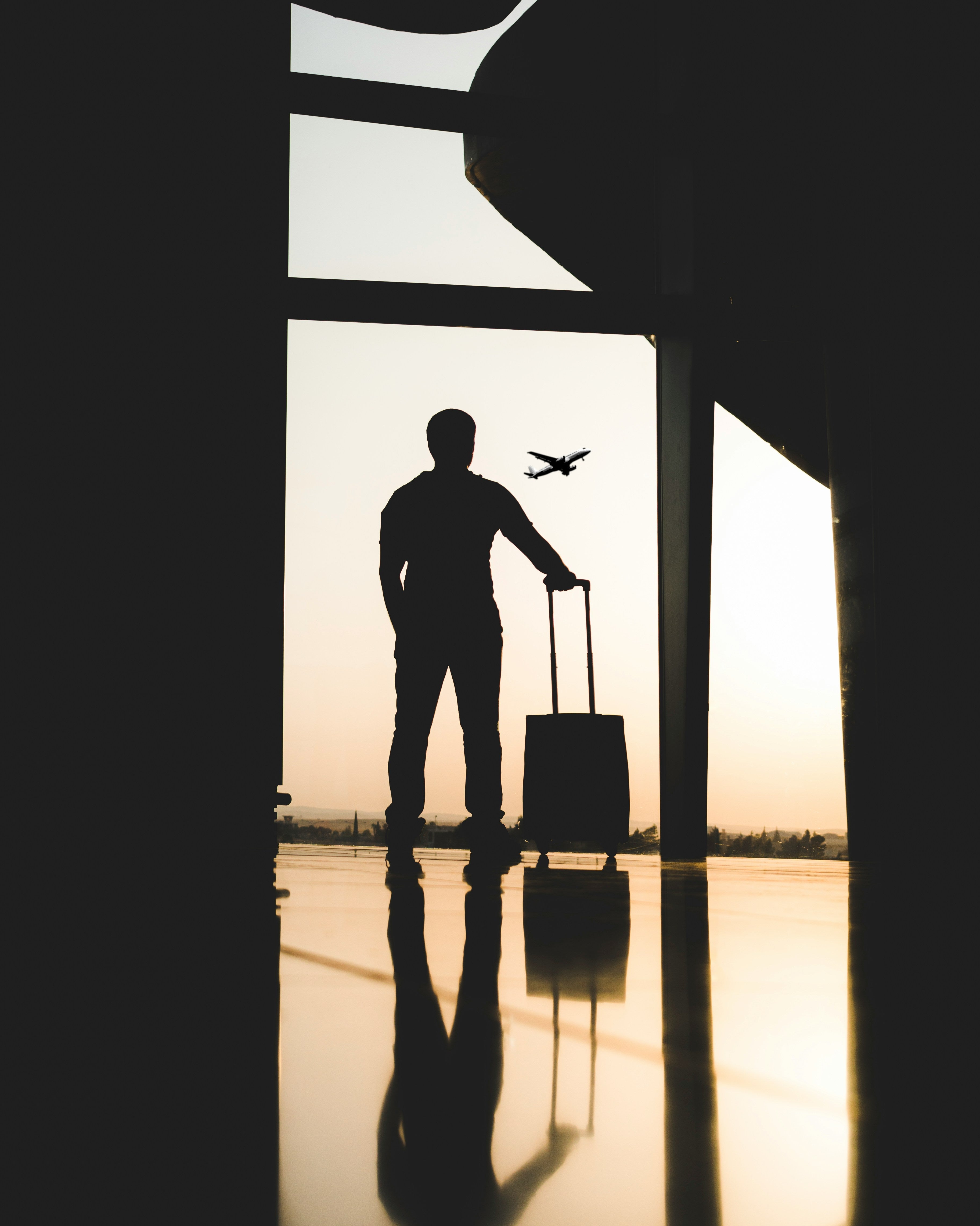 personne de dos avec sa valise à la main dans un couloir devant e couché de soleil à l'aéroport, on voit un avion décollé.